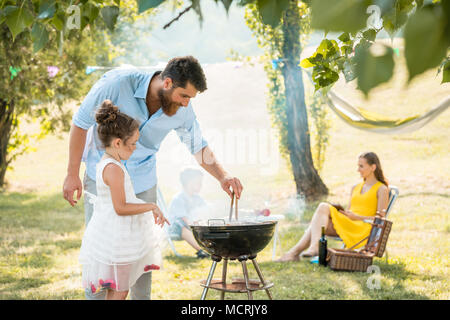 Mädchen aufpassen Vater die Zubereitung von Fleisch am Grill zubereitet, während Familie Picknick Stockfoto