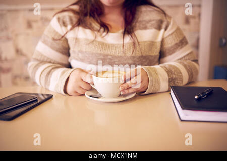 Headless Ernte eines leicht overwight Frau an einem Cafe Tabelle mit Ihrem Notizbuch und Tablet auf jeder Seite von ihr sitzen, und eine frische Tasse Kaffee Stockfoto