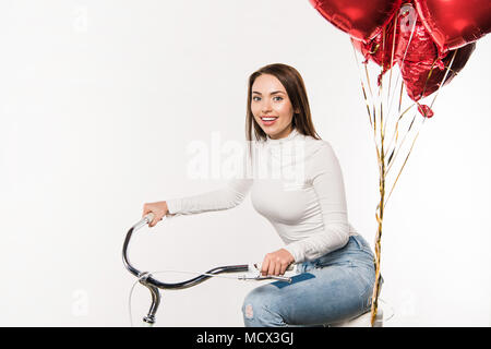 Lächelnde Frau sitzt auf Fahrrad mit roten Ballons isoliert auf weißem Stockfoto