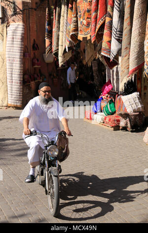 Muslimischen Mann in traditioneller Kleidung und ausgefallene Sonnenbrille auf Motorrad vorbei an einem Marktstand verkaufen Teppiche und andere Textilien in Marrakesch, Marokko Stockfoto