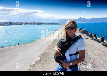 Mutter mit Baby auf Bootssteg mit Blick auf das Meer und die Berge in der Ferne, Heraklion, Griechenland Stockfoto