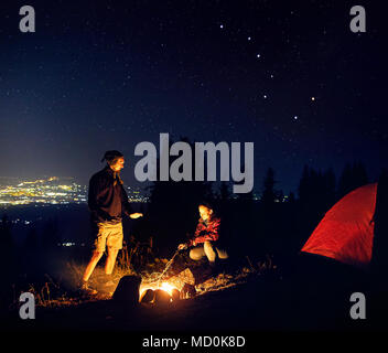 Glückliches Paar Wanderer wärmen Sie die Hände in der Nähe von Lagerfeuer unter Sternenhimmel mit Sterne und die Lichter der Stadt im Hintergrund. Stockfoto