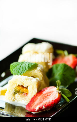 Weiße Brötchen mit Kiwi, Pfirsiche, Erdbeeren, Minze. Serverd mit Mandel Pfirsich- und Schokolade Saucen auf schwarzem Teller. Süße Nachspeise für Mädchen, Frau, Stockfoto