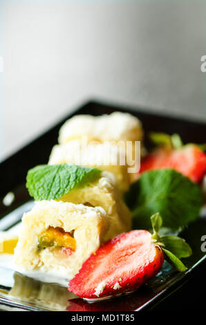 Weiße Brötchen mit Kiwi, Pfirsiche, Erdbeeren, Minze. Serverd mit Mandel Pfirsich- und Schokolade Saucen auf schwarzem Teller. Süße Nachspeise für Mädchen, Frau, Stockfoto