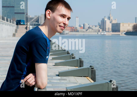 Der Kerl im blauen Hemd steht, stützte sich auf das Geländer am Ufer des Flusses, und blinzeln in die Sonne. Stockfoto