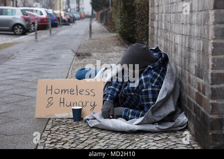 Männliche Bettler auf der Straße liegend mit Wohnungslosen und hungrig Text auf Karton Stockfoto