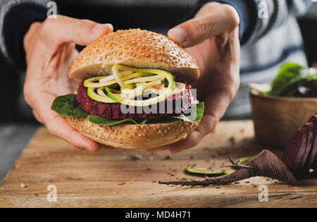 Nahaufnahme eines jungen kaukasischen Mann mit einem zuckerrüben burger Sandwich in der Hand, neben einem rustikalen Holztisch und einer in Scheiben geschnittenen Rüben Stockfoto