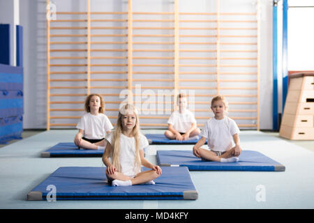 Kinder Sitzen mit gekreuzten Beinen auf blauen Matten in der Sporthalle der Schule Stockfoto