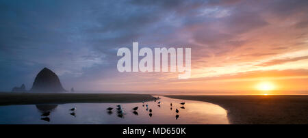 Cannon Beach bei Sonnenuntergang mit Haystack Rock, Bach und Paar mit Hund. Oregon Stockfoto