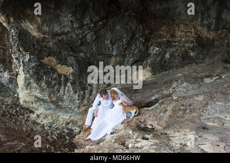 Paar Brautpaar Braut und Bräutigam Lachen und Lächeln zu einander, glücklichen und freudigen Moment. Der Mann und die Frau in der Hochzeit Kleidung sitzen auf dem Rock Hintergrund. Stockfoto