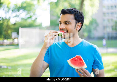 Closeup Portrait, Kerl im blauen Hemd hinunter chowing auf Wassermelone Keil außerhalb, isoliert im Freien, grünen Park Hintergrund. Organische frisches Obst Diät Stockfoto