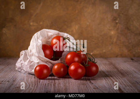 Horizontale Foto mit wenigen Zweige voller kleiner roter Kirsche Tomaten. Gemüse wird in verschlissenen zerknittertes Papier Beutel verpackt. Essen ist auf Holzbrett mit Weiß Stockfoto