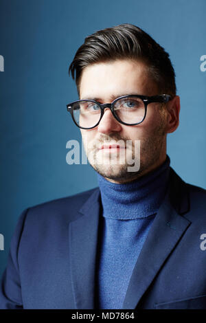 Attraktive zuversichtlich kaukasischen Mann mit Rollkragen, Jacke und Brille mit Kamera auf blauem Hintergrund Stockfoto