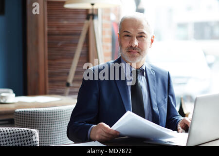 Portrait von erfolgreichen Grauhaarige kaukasischen Unternehmer mit Blick auf die Kamera beim Sitzen am Tisch mit Papieren und Laptop Stockfoto