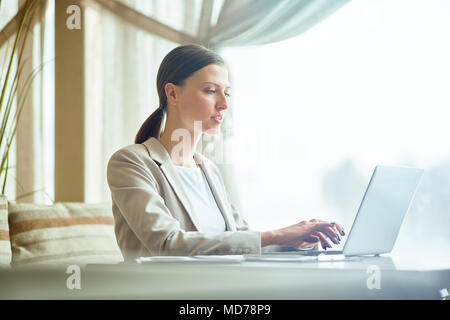 Junge elegante Kaukasischen Geschäftsfrau Tippen auf Laptop am Tisch im Restaurant sitzen und warten auf das Mittagessen Stockfoto
