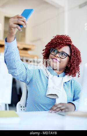 Stilvolle Afrikanische amerikanische weibliche Büroangestellte mit rotem lockigem Haar sitzt am Schreibtisch und lächelnd während der Einnahme von selfie auf dem Smartphone Stockfoto