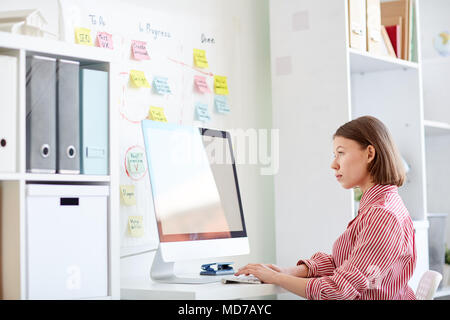Seitenansicht der jungen kaukasischen Frau in gestreiften Hemd sitzt am Schreibtisch und Arbeiten am Computer im modernen Büro Stockfoto