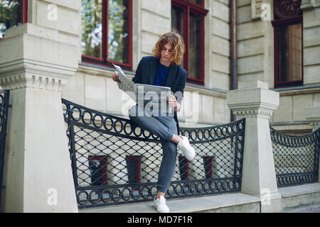 Junge rötlich, rothaarige attraktiver Mann mit lockigem Haar, eine Zeitung lesen sitzen Bein über Bein in der Nähe einer städtischen alten Gebäude. Jugend in Aktion y Stockfoto