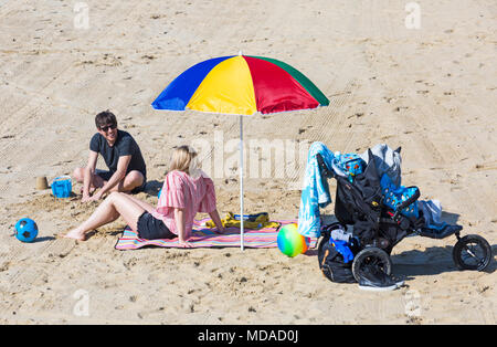 Bournemouth, Dorset, Großbritannien. 19. April 2018. UK Wetter: schön warmen sonnigen Tag in Bournemouth Stränden mit klarem blauen Himmel und Sonnenschein, ungebrochen als Besucher der seaside Leiter der wärmste Tag des Jahres so weit zu genießen. Paar am Strand entspannen unter dem bunten Sonnenschirm mit Kinderwagen Buggy durch die Seite. Credit: Carolyn Jenkins/Alamy leben Nachrichten Stockfoto