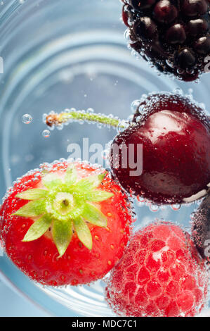 Overhead Nahaufnahme von einer Auswahl an frischem Obst, schwimmend auf der Oberseite des blau schimmernden Wasser. Mit Erdbeere, Kirsche, Himbeere und Brombeere. Stockfoto