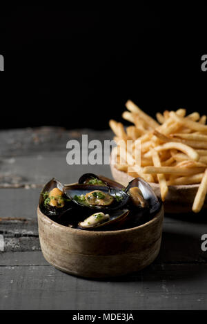 Moules - Pommes frites, Muscheln und Pommes frites, die typisch für Belgien, auf einem rustikalen Holztisch, auf einem schwarzen Hintergrund, mit einigen leeren Raum auf der Oberseite Stockfoto