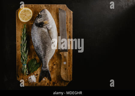 Blick von oben auf die dorado Fisch und Zitrone mit Rosmarin und Messer auf dunklen Tisch. Stockfoto