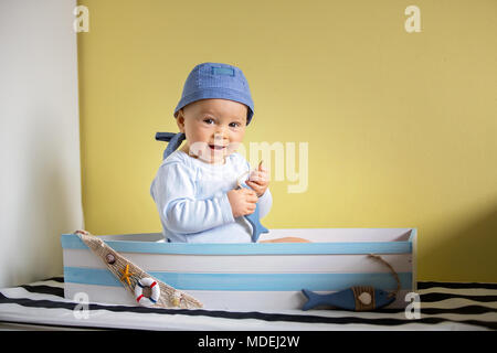 Cute Baby Boy, sitzen in einem grossen hölzernen Boot zu Hause, spielen mit Holz- fische, lächelnd Stockfoto