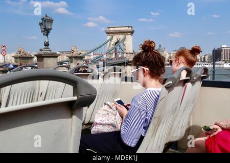 Budapest, Ungarn: Heiße hellen sonnigen Frühlingstag, April 2018. Zwei junge Mädchen auf ihren Handys auf einem oben offenen Hop-on-Hop-off Tour Bus. Stockfoto