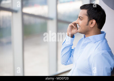 Eine Nahaufnahme Porträt einer jungen besetzt, glücklich, Lächeln business Mann im blauen Hemd, über sein Telefon, schauen aus dem Fenster, die auf Indoor gla isoliert Stockfoto