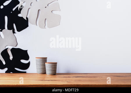 Close-up in weißer Keramik Becher mit Deckel aus Holz auf einem Schreibtisch und Blatt Schablonen Dekorationen auf eine weiße leere Wand Stockfoto