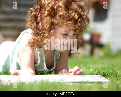 Landschaft Portrait von einem drei Jahre alten Kaukasische Mädchen mit lockigem Haar auburn im Gras an einem sonnigen Tag während Sie lesen ein Buch zur Festlegung Stockfoto