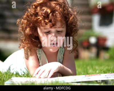 Landschaft Portrait von einem drei Jahre alten Kaukasische Mädchen mit lockigem Haar auburn im Gras an einem sonnigen Tag während Sie lesen ein Buch zur Festlegung Stockfoto