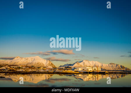 Sonnenuntergang am Henningsvær Küstenlinie mit Fabrik und riesige Berge mit Schnee bedeckt in einem wunderschönen blauen Himmel spiegelt in den See auf den Lofoten Inseln, Austvagoya Stockfoto