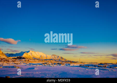 Sonnenuntergang am Henningsvær Küstenlinie mit riesige Berge mit Schnee bedeckt in einem wunderschönen blauen Himmel auf den Lofoten Inseln, Austvagoya Stockfoto