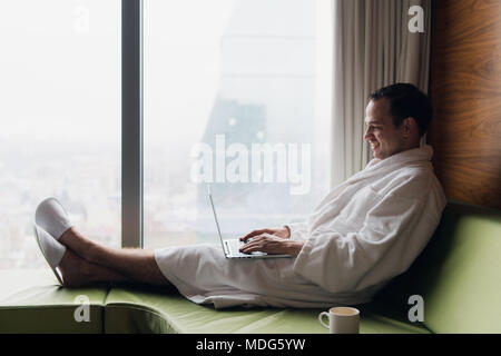 Junge lächelnde Geschäftsmanns, der auf dem Laptop Computer tragen weiße Bademantel saß in der Nähe der Fenster mit Tasse Kaffee bei Sonnenaufgang Blick auf die Stadt. Motivation Konzept Stockfoto