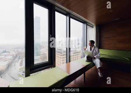 Unternehmer zu Hause oder in der Reise. Seitenansicht Portrait von hübscher junger Geschäftsmann sitzt auf dem Bett und mit Laptop. Panoramafenster mit schönen Sonnenaufgang Stadt Landschaft im Hintergrund Stockfoto