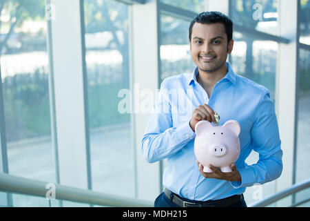 Closeup Portrait glücklich lächelnd Geschäftsmann, setzen, Geld in rosa Sparschwein, isoliert im Innenbereich Büro Hintergrund. Finanzielle Einsparungen, smart inv Stockfoto