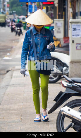 Eine vietnamesische Frau trug eine kegelförmige Hut, Jeans Jacke und grüne Hose zu Fuß auf dem Gehweg auf einer Straße in Ho Chi Minh City, Vietnam. Stockfoto