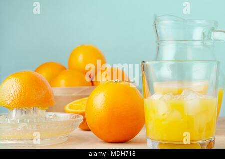 Manuelle Saft pressen Prozess mit ganzen und geschnittenen Orangen, squeezer, Kanne und halb gefüllten Glas mit Eiswürfel. Holz Tisch mit aqua Hintergrund. Stockfoto