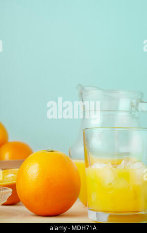 Manuelle Saft pressen Prozess mit ganzen und geschnittenen Orangen, Kanne und halb gefüllten Glas mit Eiswürfel. Holz Tisch mit aqua Hintergrund. Stockfoto