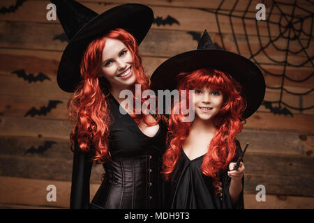 Schönen weißen Mutter und ihre Tochter mit langen roten Haaren in Hexe Kostüme und Zauberstab feiern Halloween posiert mit über Fledermäuse und Spinnennetz auf Holz- studio Hintergrund Stockfoto