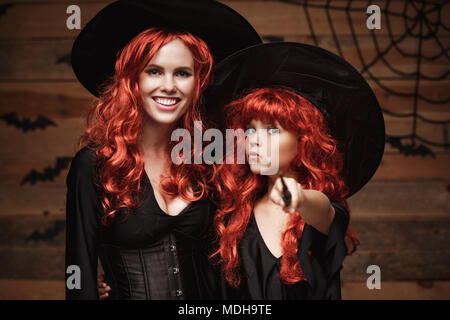 Schönen weißen Mutter und ihre Tochter mit langen roten Haaren in Hexe Kostüme und Zauberstab feiern Halloween posiert mit über Fledermäuse und Spinnennetz auf Holz- studio Hintergrund. Stockfoto