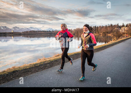 Zwei junge Frauen, die auf einem Wanderweg am Rand des Wassers mit Bergen in der Ferne, Anchorage, Alaska, Vereinigte Staaten von Amerika Stockfoto