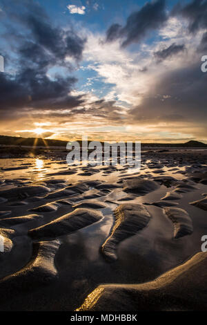 Nassen Wellen in den Sand am Strand bei Sonnenuntergang mit einem Sunburst über Silhouetted Hügel in der Ferne; Embleton, Northumberland, England Stockfoto