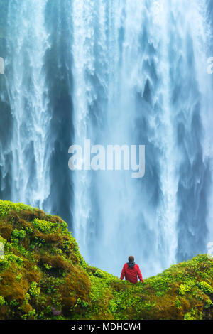 Mann sitzt auf einem bemoosten Felsen an der Basis der Skogafoss Wasserfall ; Island Stockfoto