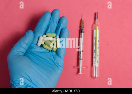 Sortiment von Pillen, Tabletten und Kapseln auf Hand in Blau Handschuh gegen Rosa Hintergrund, Spritze und Thermometer. Stockfoto