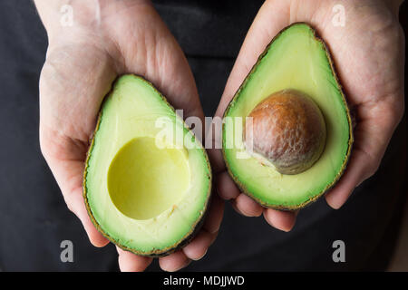 Junge kaukasier Frau in Schwarz Schürze Holding in den Händen reif Schön halbiert Organische australische Avocado mit Grube. Gesunder Lebensstil Vegan Vitamin E Öl Stockfoto