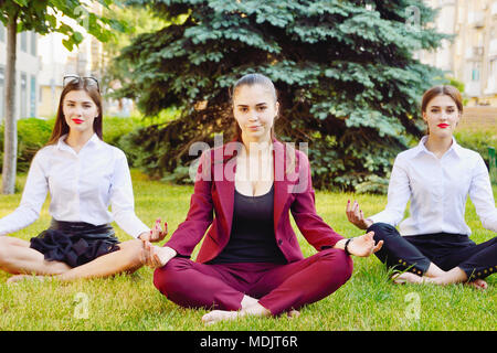 Büro Yoga. Drei junge Mädchen im Lotussitz sitzen auf grünem Gras und Meditieren. Freizeit der Mitarbeiter des Amtes. Gesunder Lebensstil Stockfoto