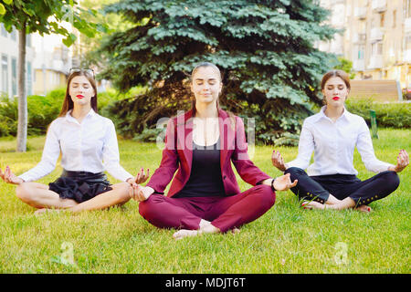 Büro Yoga. Drei junge Mädchen im Lotussitz sitzen auf grünem Gras und Meditieren. Freizeit der Mitarbeiter des Amtes. Gesunder Lebensstil Stockfoto