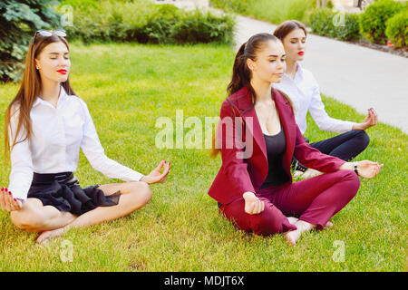 Büro Yoga. Drei junge Mädchen im Lotussitz sitzen auf grünem Gras und Meditieren. Freizeit der Mitarbeiter des Amtes. Gesunder Lebensstil Stockfoto